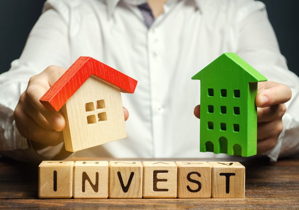 A person in a white shirt holding two wooden house models, one resembling a small home with a red roof and the other a green apartment building, with wooden blocks spelling "INVEST" in front.