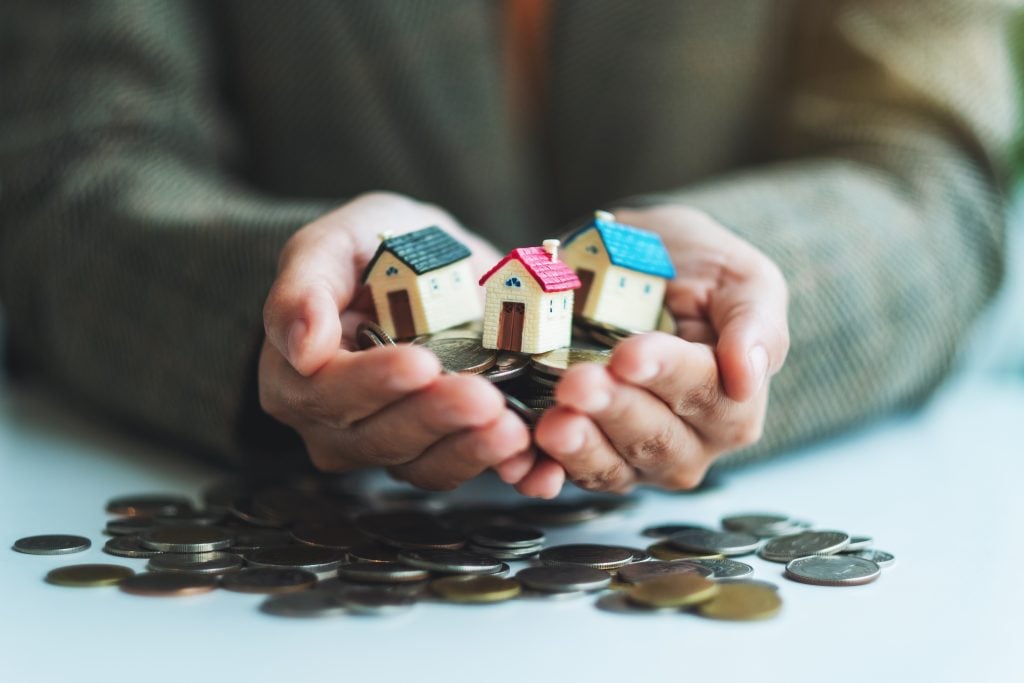 A person’s hands holding small wooden house models over a pile of coins, illustrating property investment and financial security.