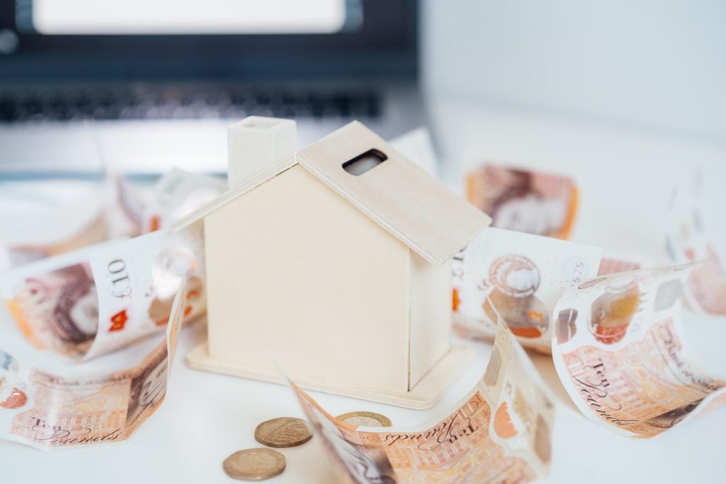 A wooden house model resembling a piggy bank surrounded by British banknotes and coins, symbolising savings, homeownership, or mortgage planning.