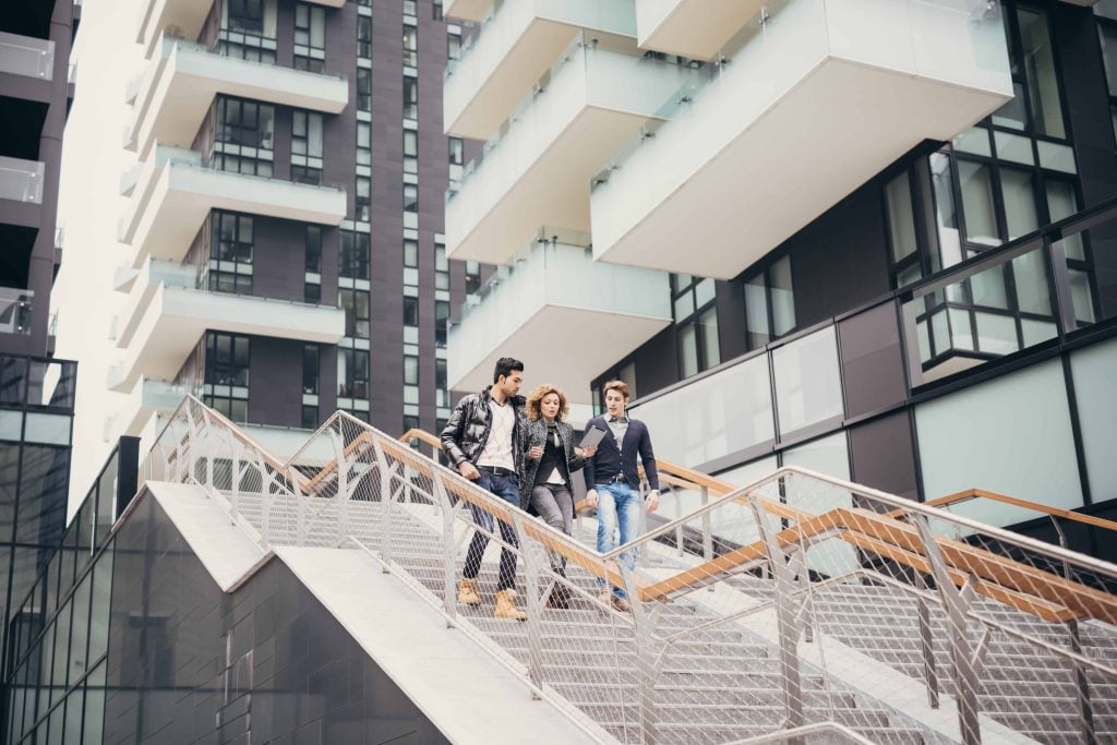 Group of three young students walking down stairs outside a modern student accomodation, looking at a digital tablet.