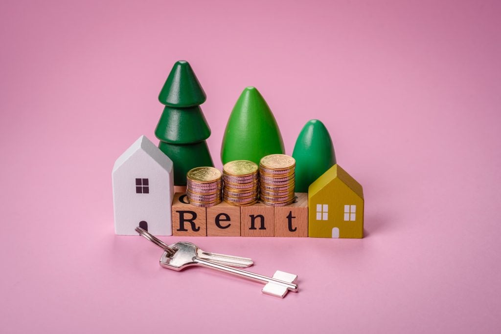 Mini houses, stacked coins, and ‘rent’ blocks with keys on pink background.