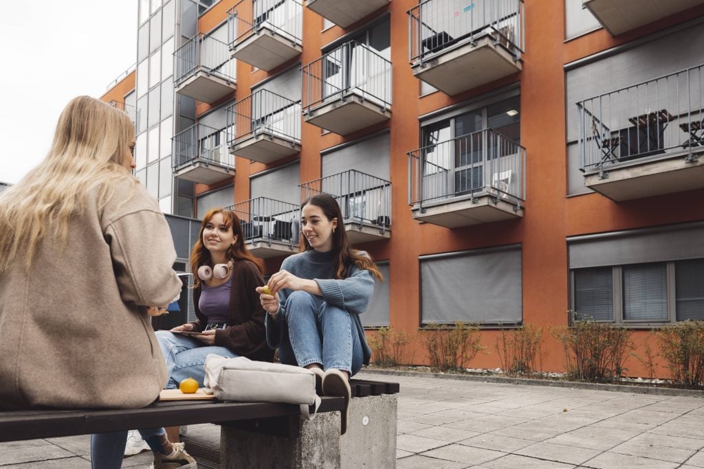 Three female students socialising outside orange student flats with balconies.