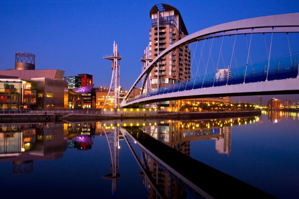 Millennium Bridge and illuminated buildings at Salford Quays, Manchester, reflected in calm evening water.