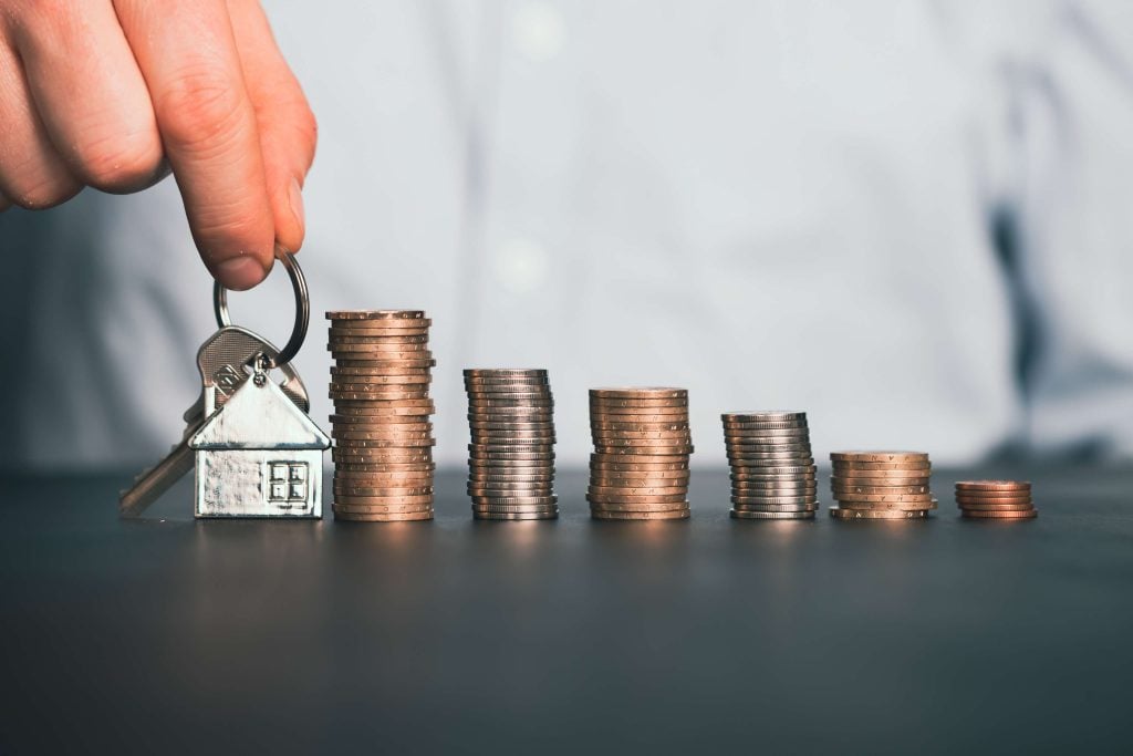 Hand holding house-shaped keychain next to rising stacks of coins, representing real estate savings or investment.