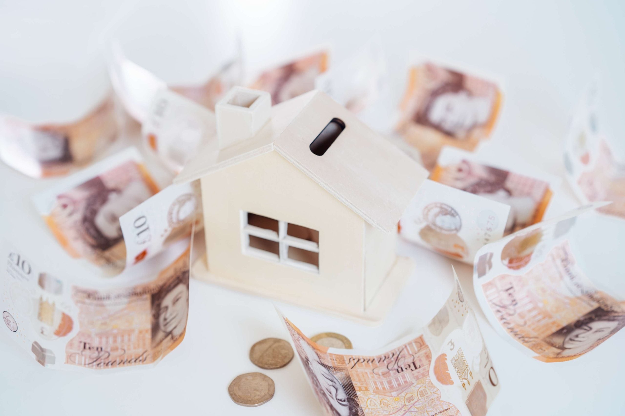 Wooden house piggy bank surrounded by British pound notes and coins, representing UK housing savings or investment.