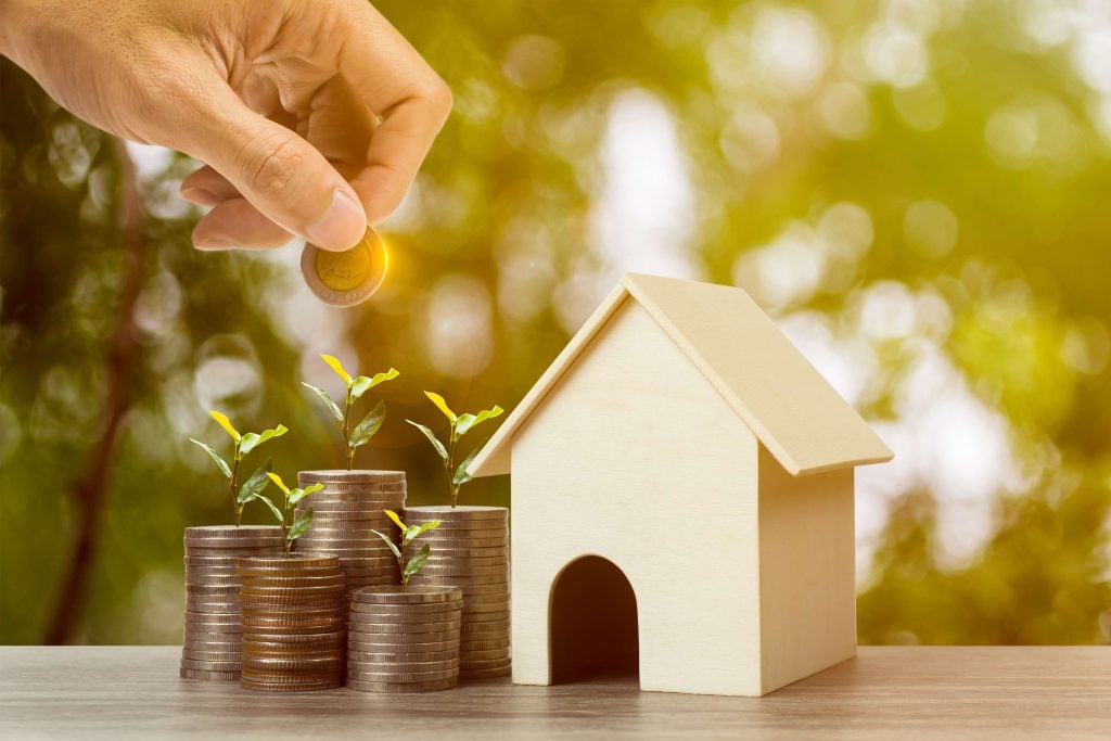 Hand placing coin near stacked money and house model.