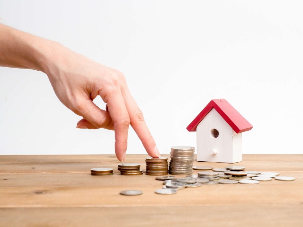 Hand walking on stacked coins towards small house model.