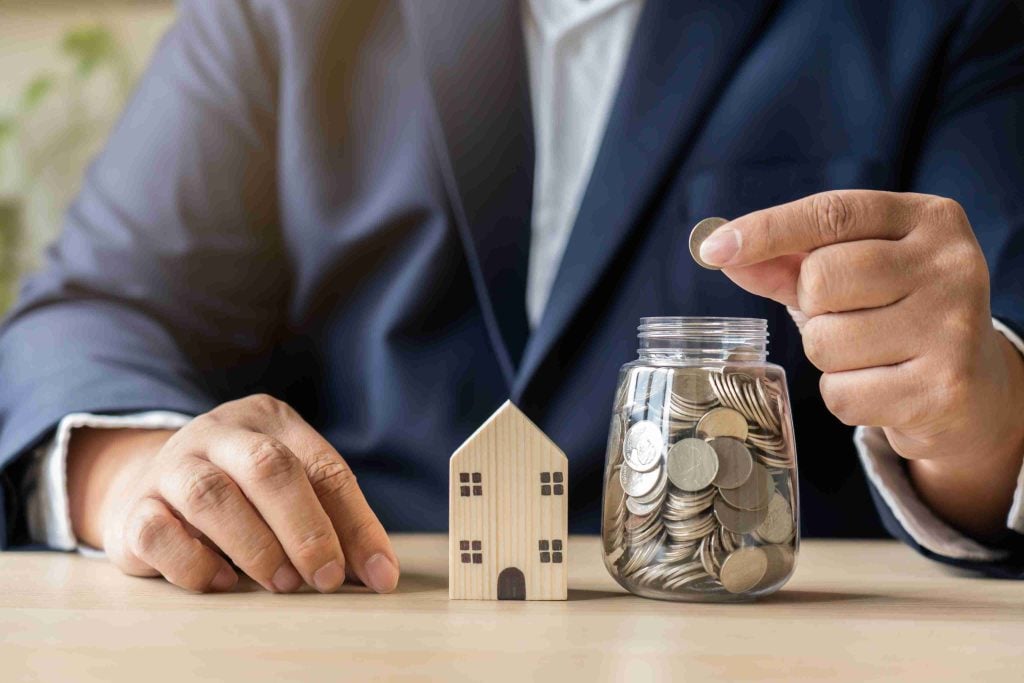 Property investor saving rental income in a coin jar next to a wooden model house, illustrating the financial returns of hands-free property investment.
