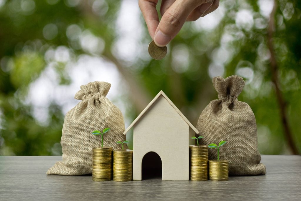 Hand adding a coin to rising stacks beside a model house, illustrating property investment growth