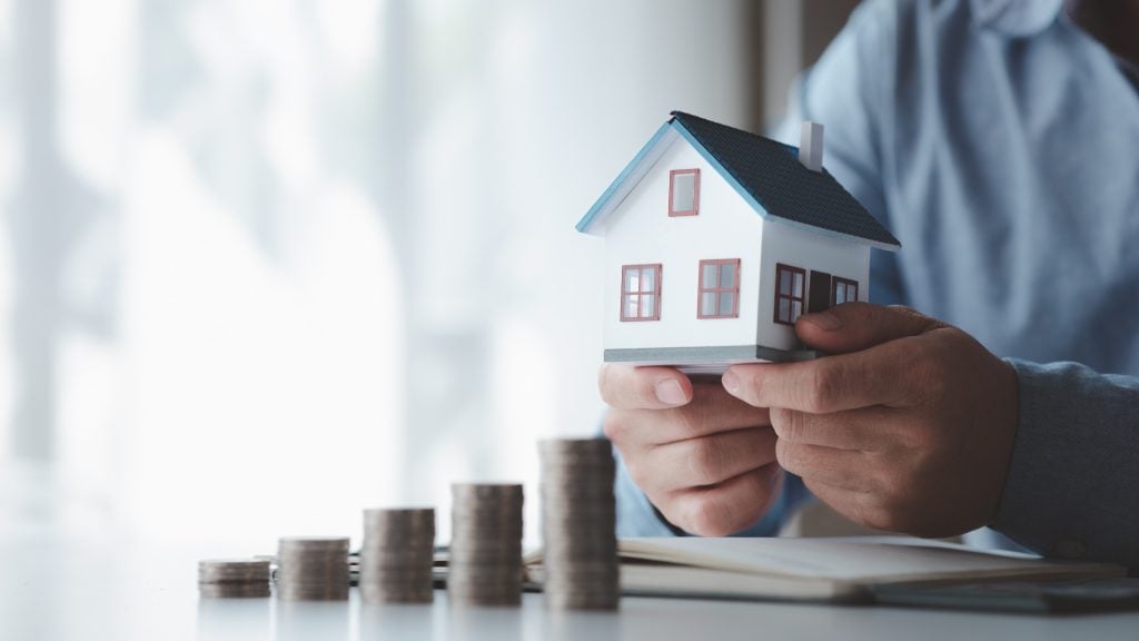 Person holding a model house above ascending coin stacks, showing long-term property investment returns